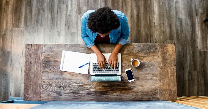 Overhead view of woman typing on laptop with coffee and notepad next to her