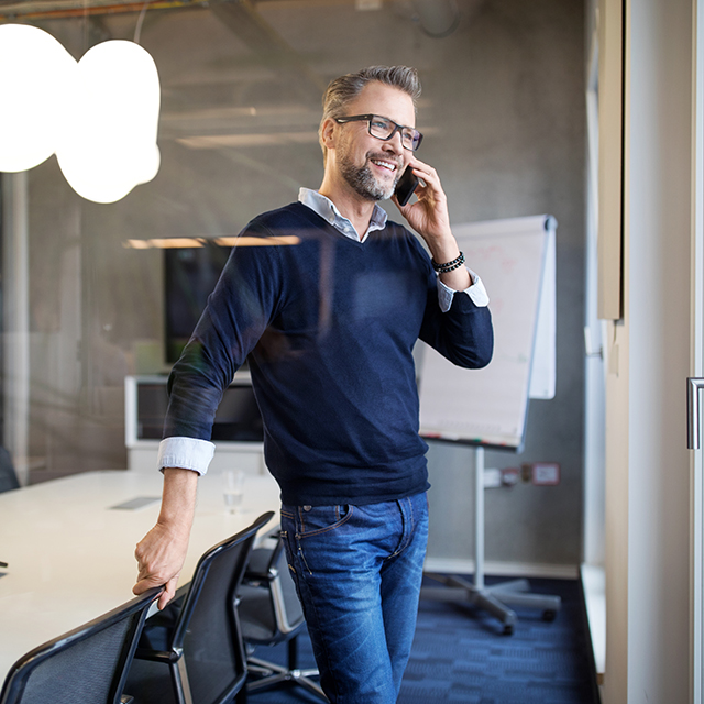 A professional standing in a modern conference room, leaning on a chair while talking on a phone near a table and whiteboard.