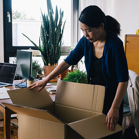 A person unpacking a large cardboard box in a home office filled with plants, a desk, and computer equipment near a window.
