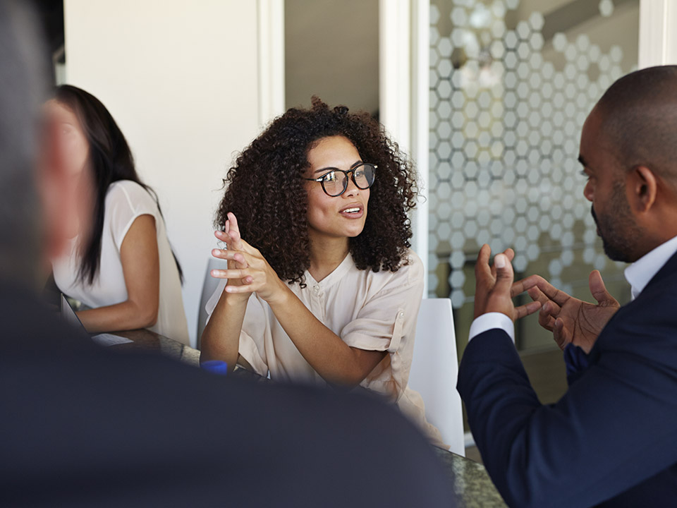 Two professionals seated at a conference table engaged in a discussion, gesturing with their hands as they talk in a modern office setting with glass walls and a patterned design in the background.