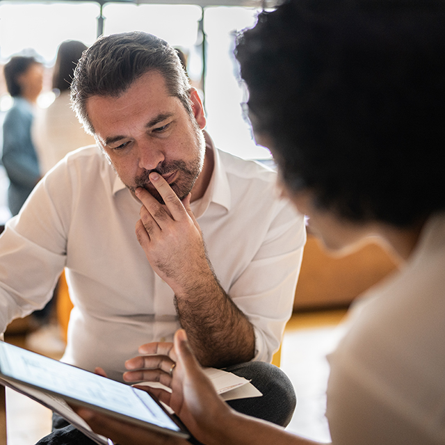 Two professionals seated and engaged in conversation, reviewing information on a tablet in a collaborative office environment.