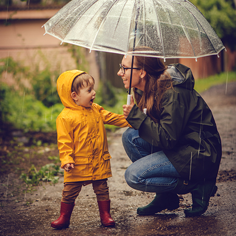 An adult crouches to speak with a young child wearing a raincoat, both standing on a wet path as rain falls and an umbrella is held overhead.