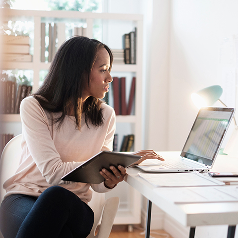 A person working at a desk with a laptop and tablet, seated in a well‑lit home office with bookshelves in the background.