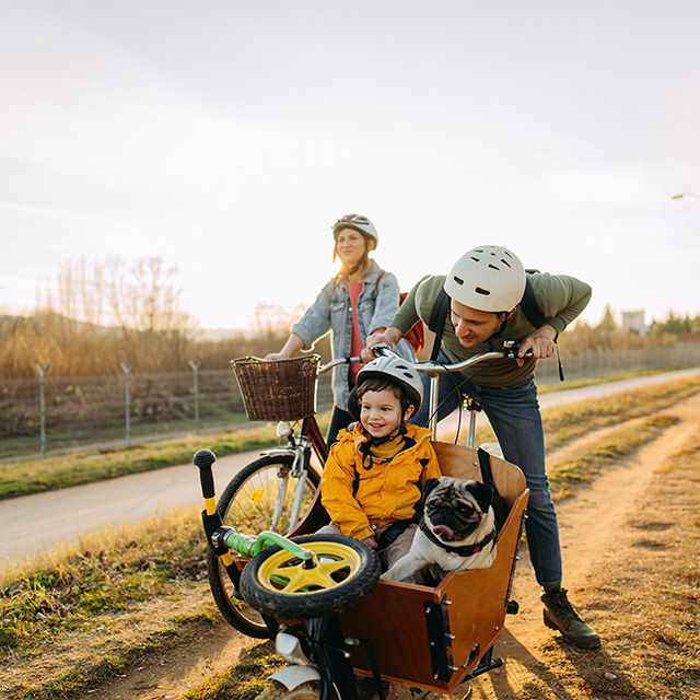A family cycling together along a dirt path, with one adult pushing a cargo bike carrying a child and a small dog, while another adult rides alongside on a bicycle in a rural setting.