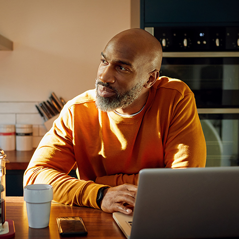 A person seated at a kitchen table working on a laptop, with a coffee mug, phone, and kitchen appliances in the background.
