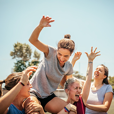 A group of people outdoors lifting and supporting one person in a playful, team‑building activity under a clear sky.