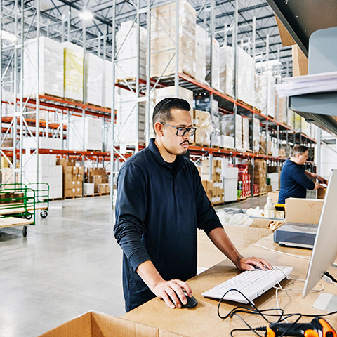 A warehouse worker standing at a workstation, using a computer to manage inventory among tall shelves stacked with boxes.